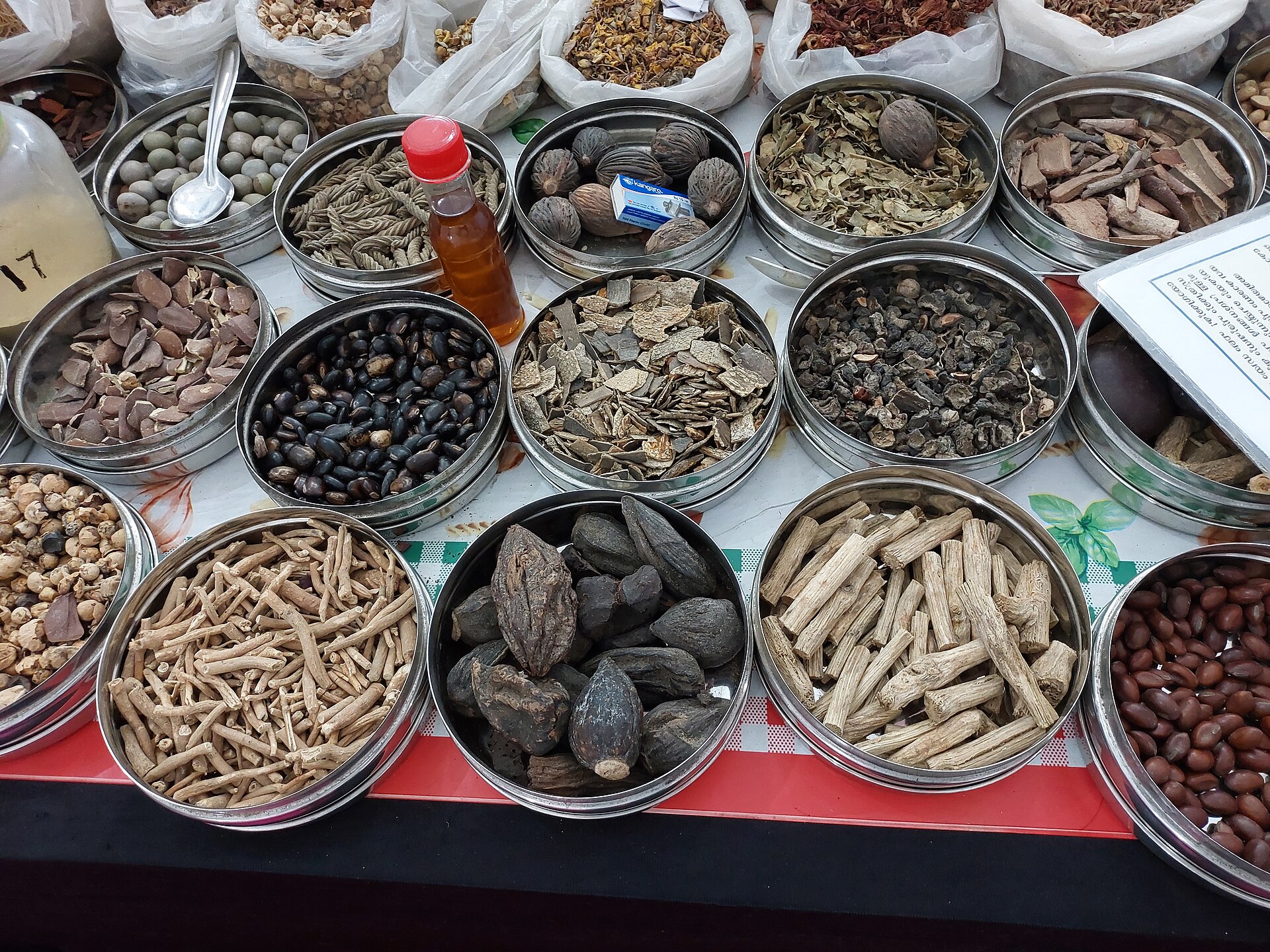 Steel bowls of dried Ayurvedic roots, barks, seeds and leaves laid out at a herbal medicine stall.